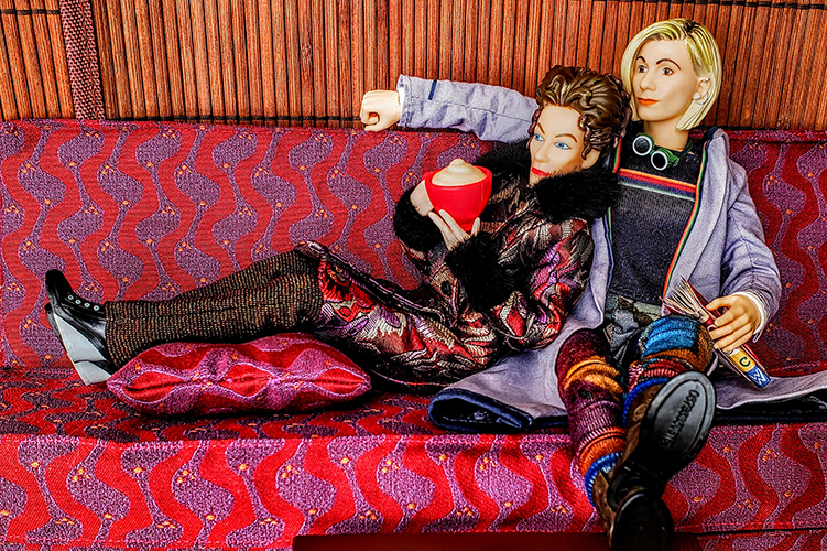 Action figures sit cozily on a couch, Missy leaning against the Thirteenth Doctor.